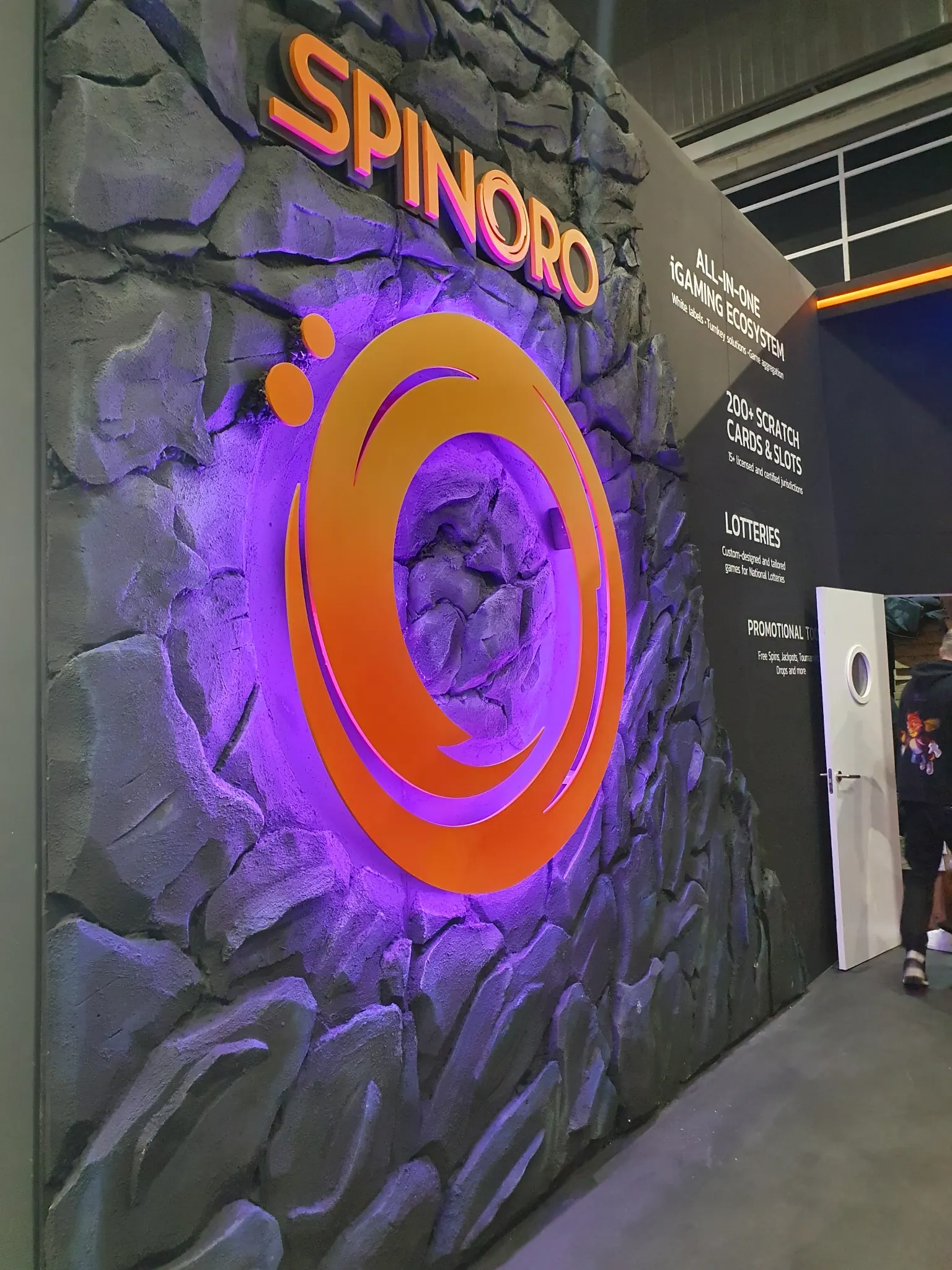Sculptural rock-effect wall with violet lighting and orange Spinoro logo, built for Anakatech / Spinoro at ICE Barcelona 2025